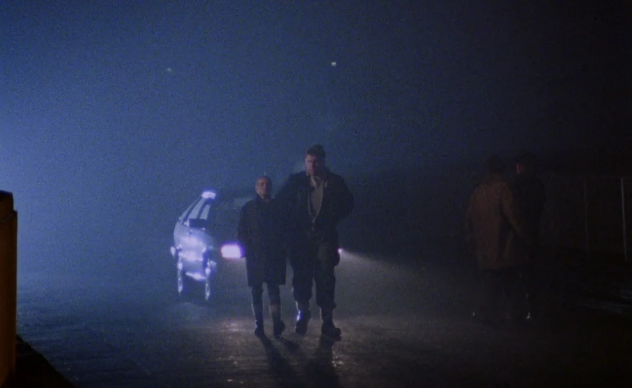 My Beautiful Laundrette (1985) movie still: wide — An open asphalt surface at night is filled with thick, low-lying blue fog. Two men walk…