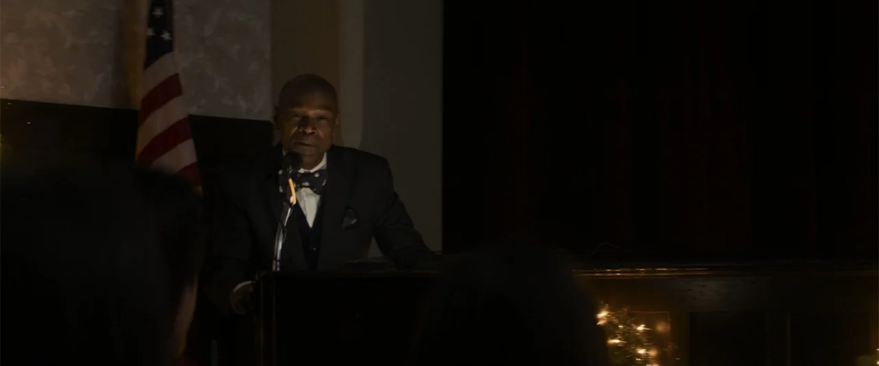 Holiday Harmony (2022) movie still: medium — A dark indoor space features a man standing at a podium near a flag. The man speaks into a microphone…