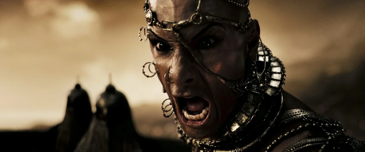 300 (2006) movie still: closeup — A bald man with intricate piercings and metal jewelry yells on a battlefield. Two blurred soldiers stand in the…