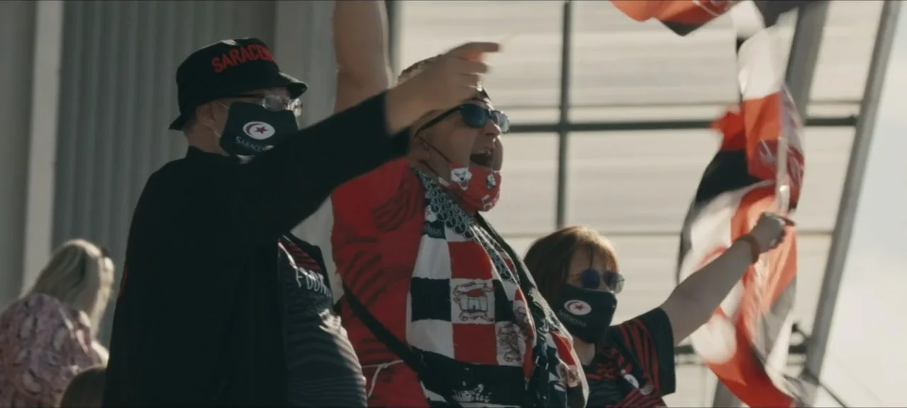 No Woman No Try (2022) movie still: medium — A group of fans wearing red and black jerseys sit in a stadium seating area. They are cheering and…