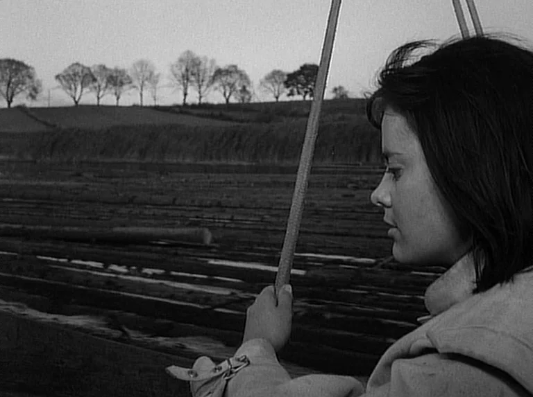 Knife In The Water (1962) movie still: medium — A dark, wood-planked structure extends into a rural landscape with distant trees. A woman in a light…