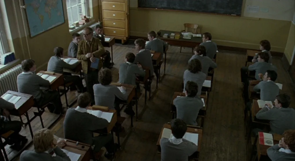 Breakfast On Pluto (2005) movie still: wide — An old school room features wooden desks, yellow walls, and a large wall map. A teacher in a brown…