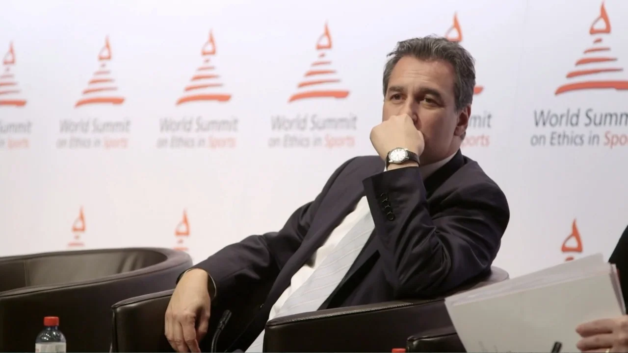 The FIFA Family: A Love Story (2017) movie still: medium — A man sits in a leather armchair in front of a white wall with branding. He is listening…