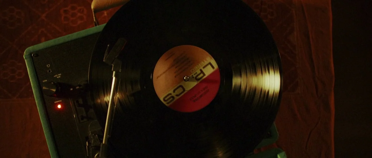 Depraved (2019) movie still: closeup — A portable turquoise vinyl record player rests on a patterned fabric surface. A black record spins on the…
