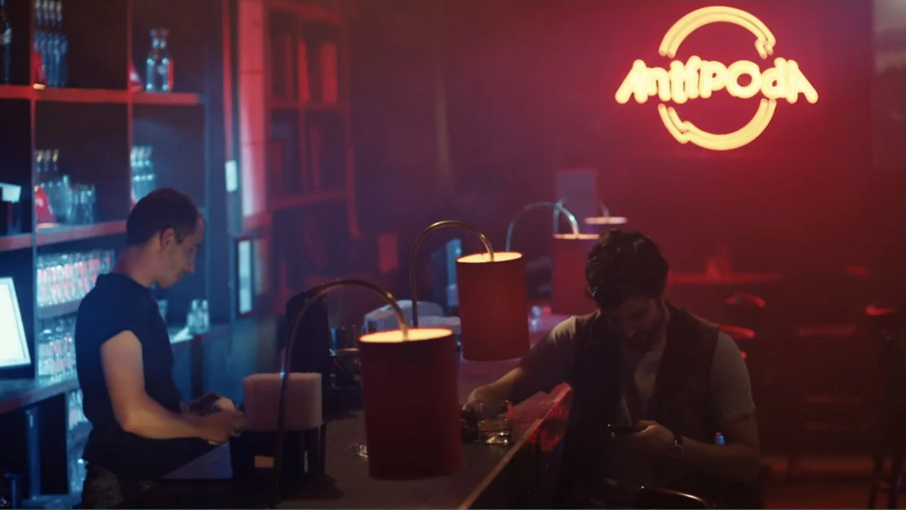 Alice in Marialand (2014) movie still: medium — A dimly lit bar with red neon signage reading Antipoda on the back wall. Two men stand at the bar…