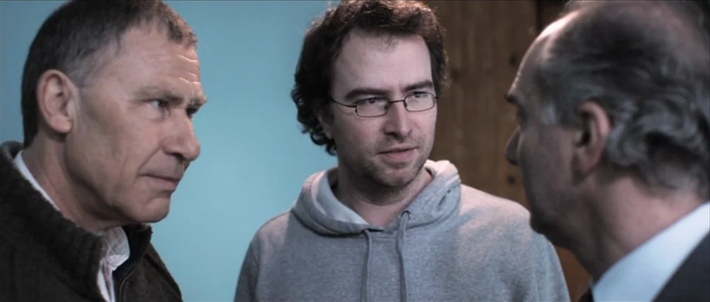 Down Terrace (2009) movie still: medium — Three men stand close together in a plain room with a pale blue wall. The central man in a hoodie looks…