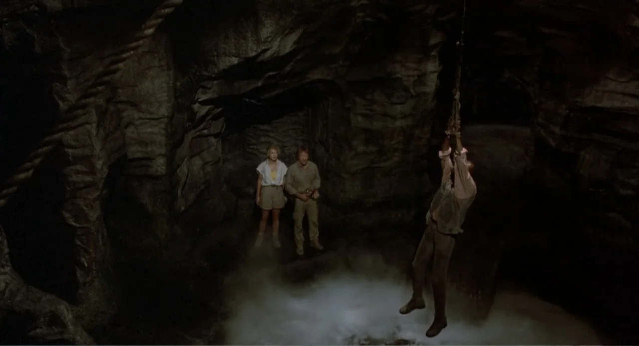 Firewalker (1986) movie still: wide — A dark rocky cavern contains a deep chasm with mist rising from below. A man hangs suspended by a rope while…