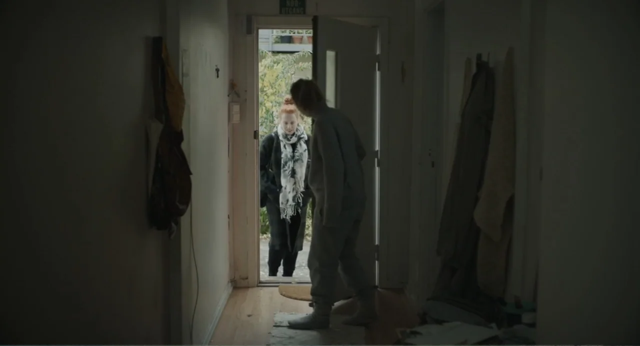 Ninjababy (2021) movie still: medium — A narrow apartment entryway features white walls, hanging coats, and an open wooden doorway. One person…