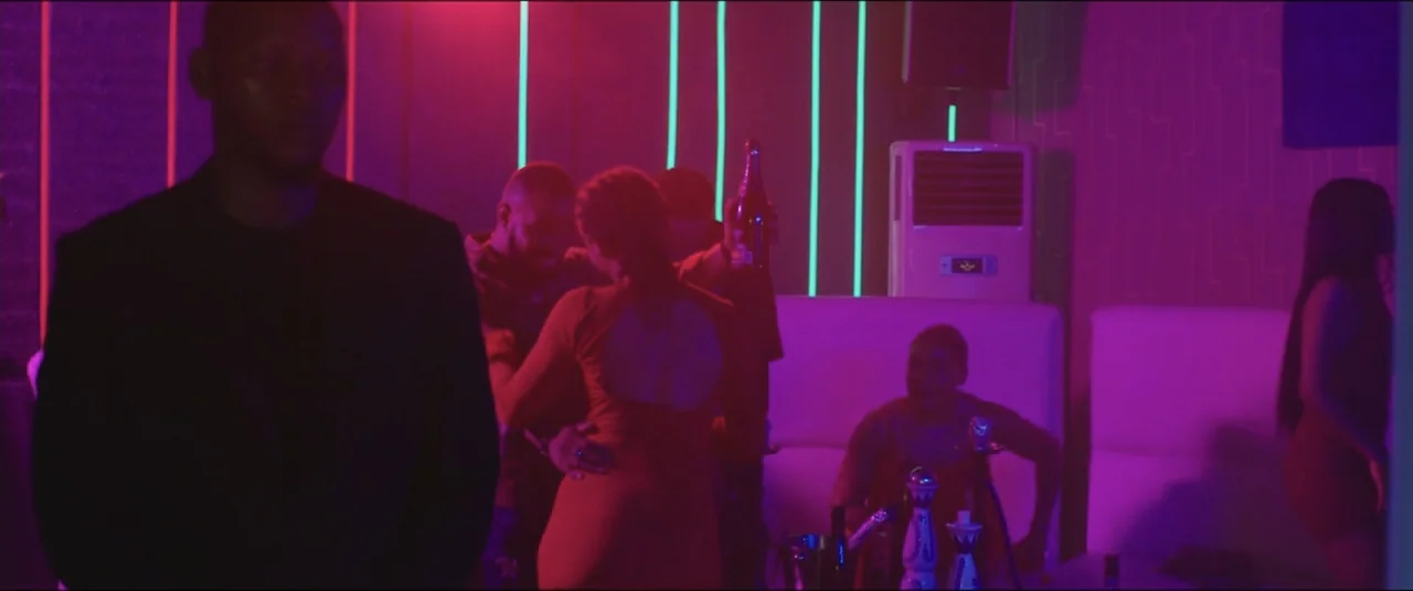 Saving Onome (2024) movie still: medium — A dark, neon-lit nightclub interior with white booths and vertical glowing wall strips. A man stands in…