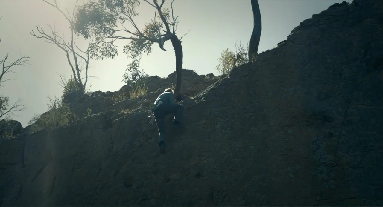 Ice Road: Vengeance (2025) movie still: wide — A rugged rock face stands under a bright sun with a single tree above. A person in grey clothing is…