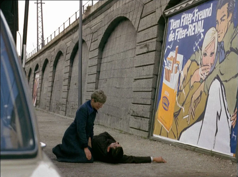 Satan’s Brew (1976) movie still: fullbody — A stone viaduct wall supports a large vintage cigarette advertisement beside a gravel path. A woman…