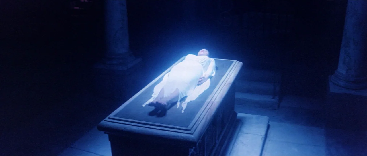Lifeforce (1985) movie still: wide — A dim underground crypt features stone pillars and a large central elevated tomb structure. A woman in a white…