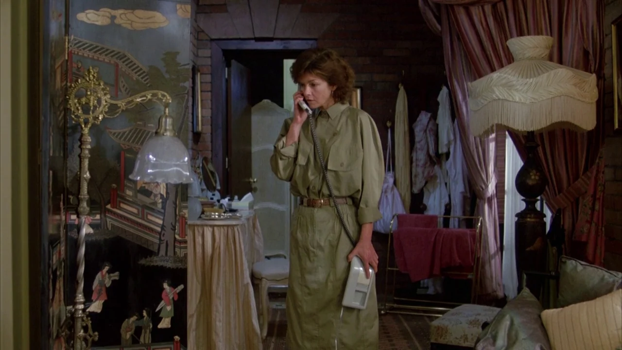 Dead Ringers (1988) movie still: medium — A room with brick walls features an oriental screen and a tall fringed lamp. A woman in a long olive dress…