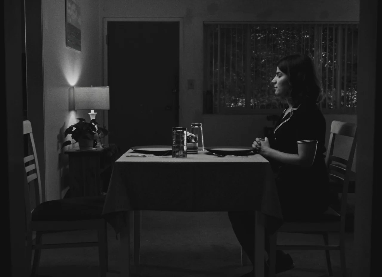 Fremont (2023) movie still: medium — A dimly lit room features a table set for two with plates and glasses. A young woman sits alone at the table…