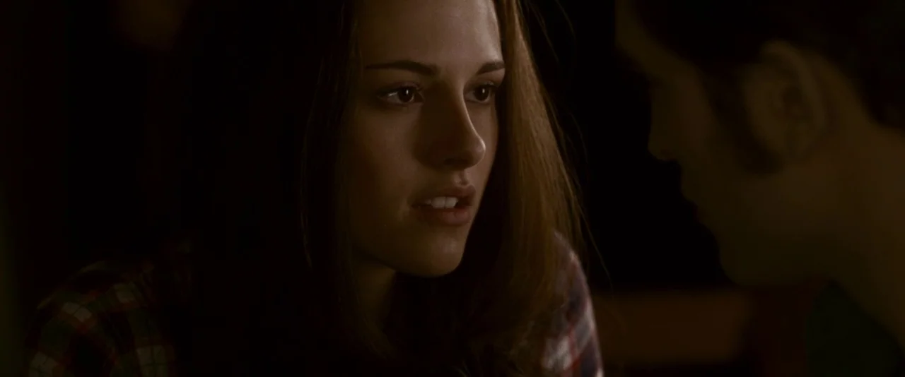 The Twilight Saga: Eclipse (2010) movie still: closeup — A dark indoor space with a dimly lit booth setting. A young woman looks at someone seated…