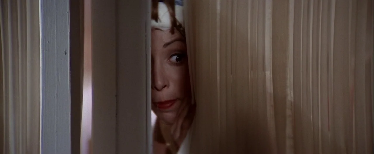 Grease (1978) movie still: closeup — A woman looks out from behind vertical window blinds in a home interior. She is peeking through the narrow…