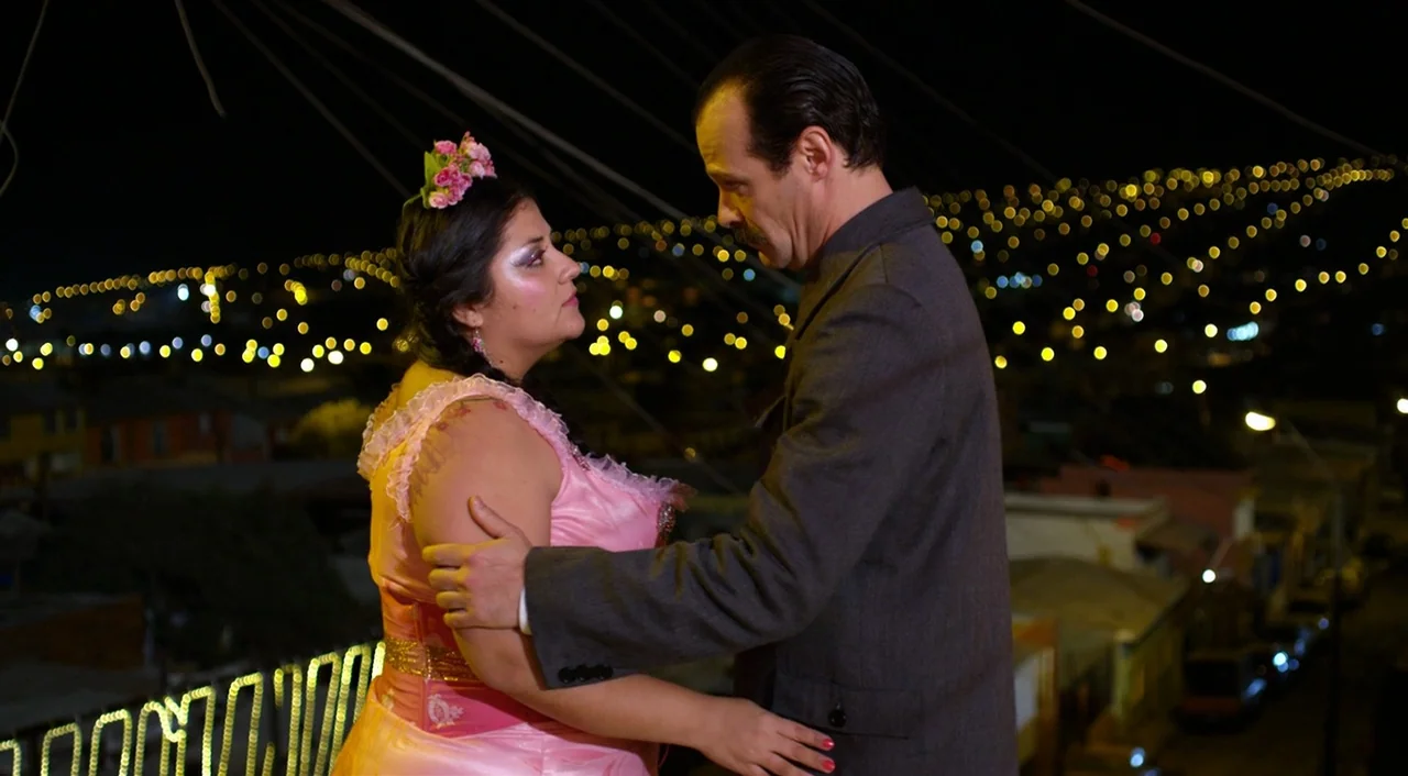 Dance of Reality (2013) movie still: medium — A dark outdoor balcony overlooks a hillside covered in golden nighttime city lights. A woman in a pink…
