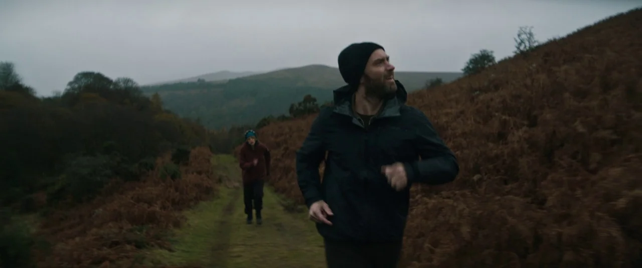 The Rhythm Section (2020) movie still: medium — A muddy trail cuts through a landscape of rolling brown hills and sparse shrubs. Two men run toward…