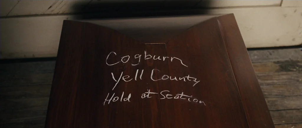 True Grit (2010) movie still: closeup — A wooden crate sits on a rough wooden floorboards in a dimly lit space. White chalk is written on the lid…