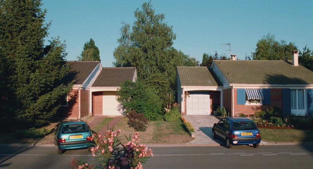 Ma Vie En Rose (1997) movie still: wide — Two adjacent red brick suburban houses with tiled roofs stand along a paved street. A teal car and a blue…