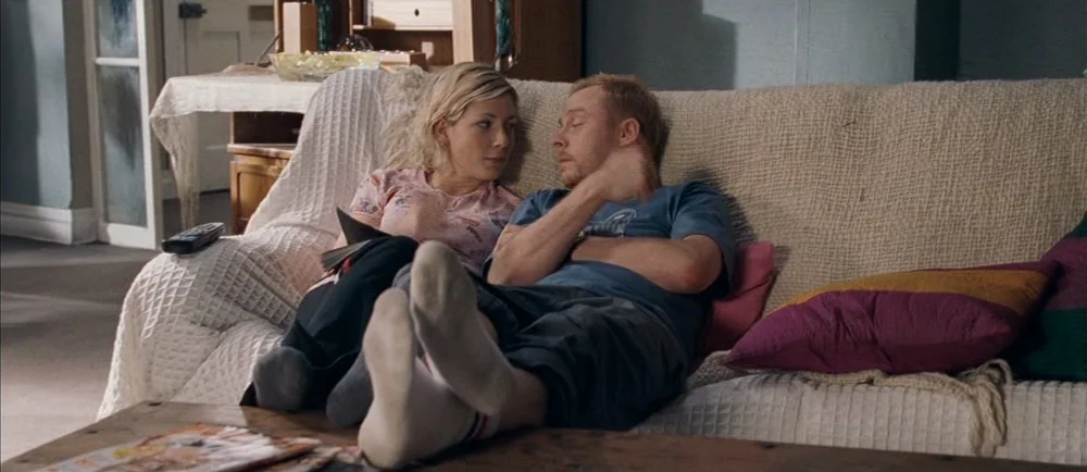 Shaun Of The Dead (2004) movie still: medium — A furnished living room with a beige textured sofa featuring various throw pillows. A couple lounges…