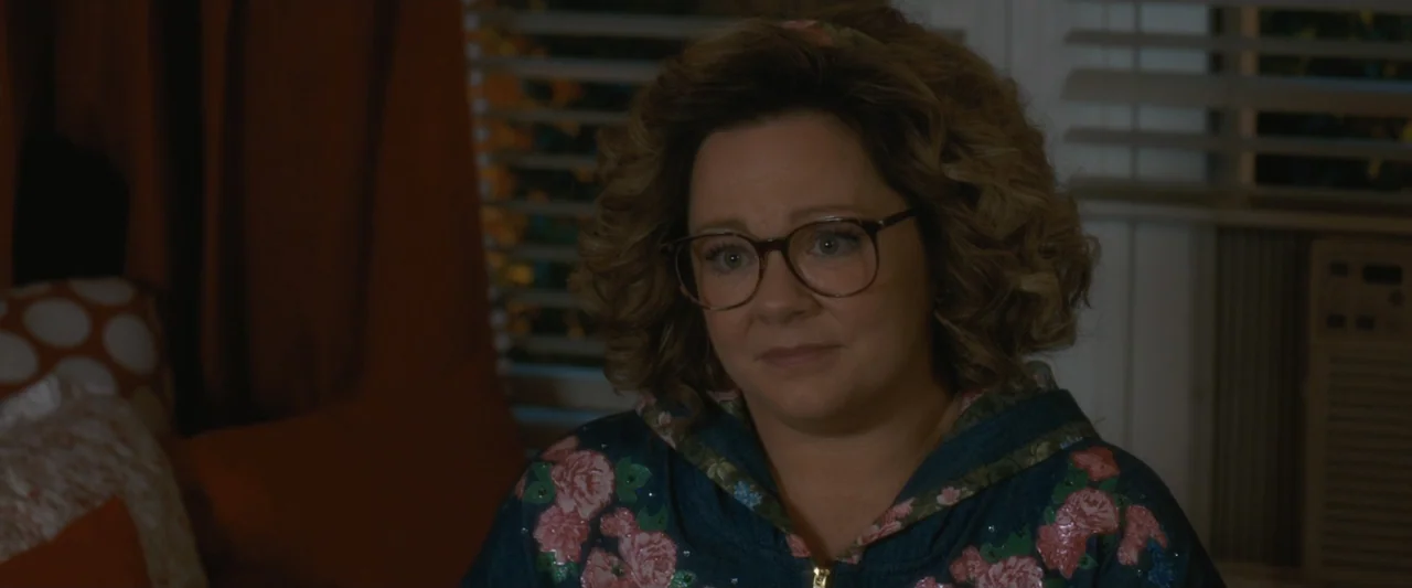 Life of the Party (2018) movie still: closeup — A dimly lit room contains horizontal window blinds and patterned orange pillows. A woman wearing…