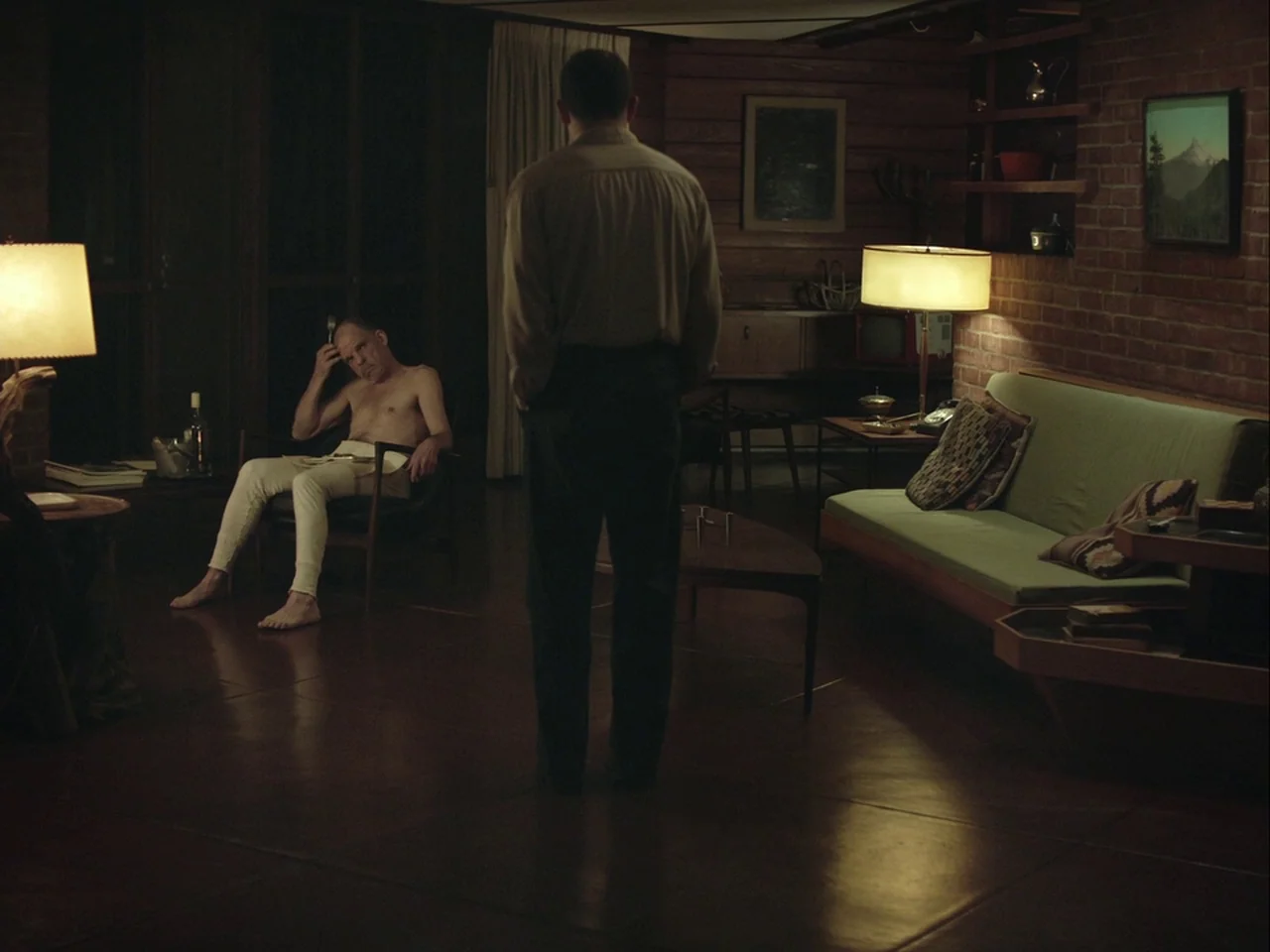 The Mountain (2018) movie still: medium — A 1970s living room features wood-paneled walls and a brick fireplace section. One man stands with his…