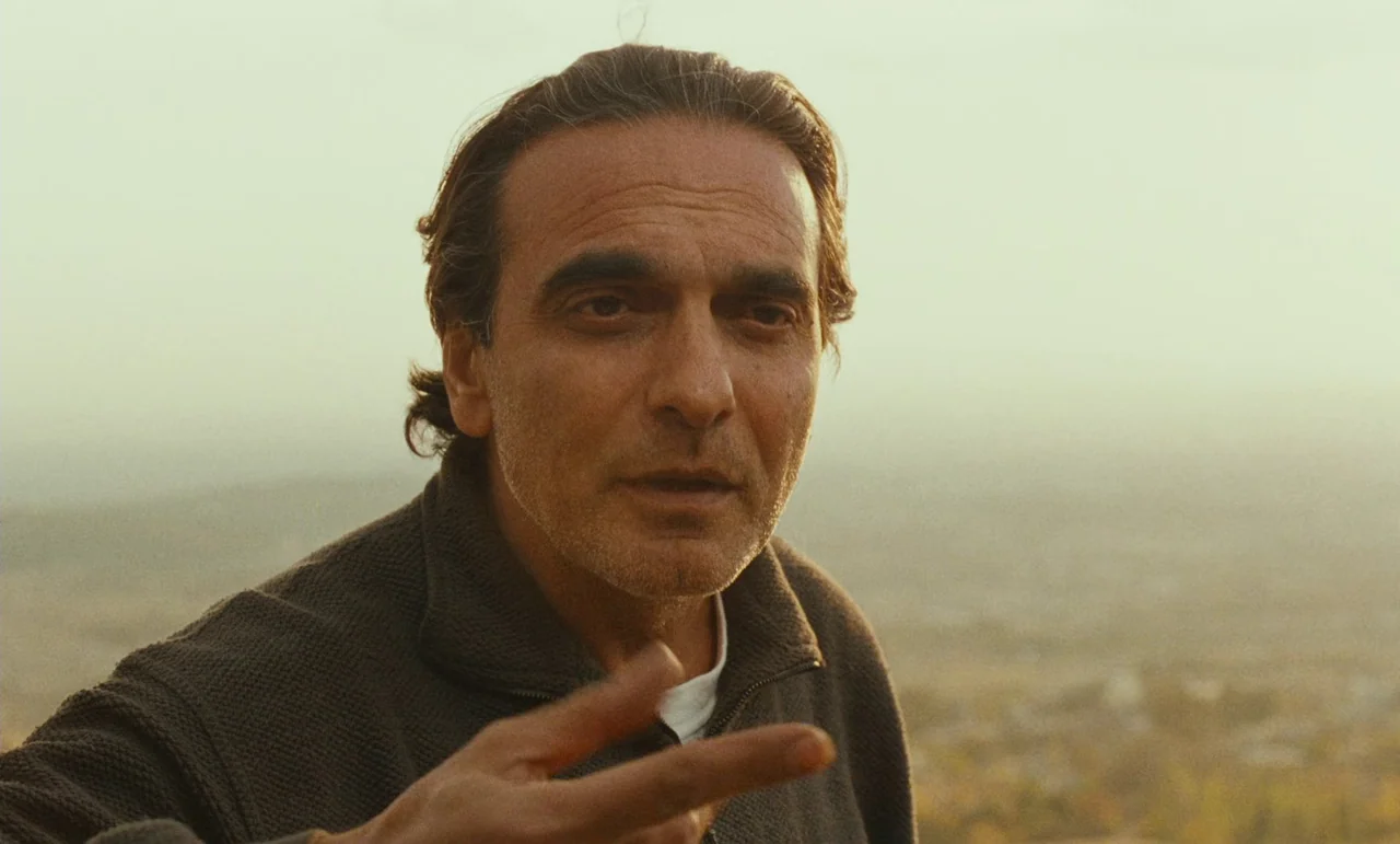 Taste of Cherry (1997) movie still: closeup — A man stands outdoors in a landscape with soft, out-of-focus earth tones. The man talks while…