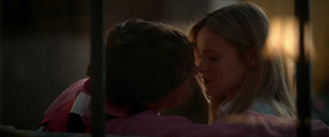 Love Me Love Me (2026) movie still: closeup — A dimly lit room interior features two young people positioned face to face closely. The young man…