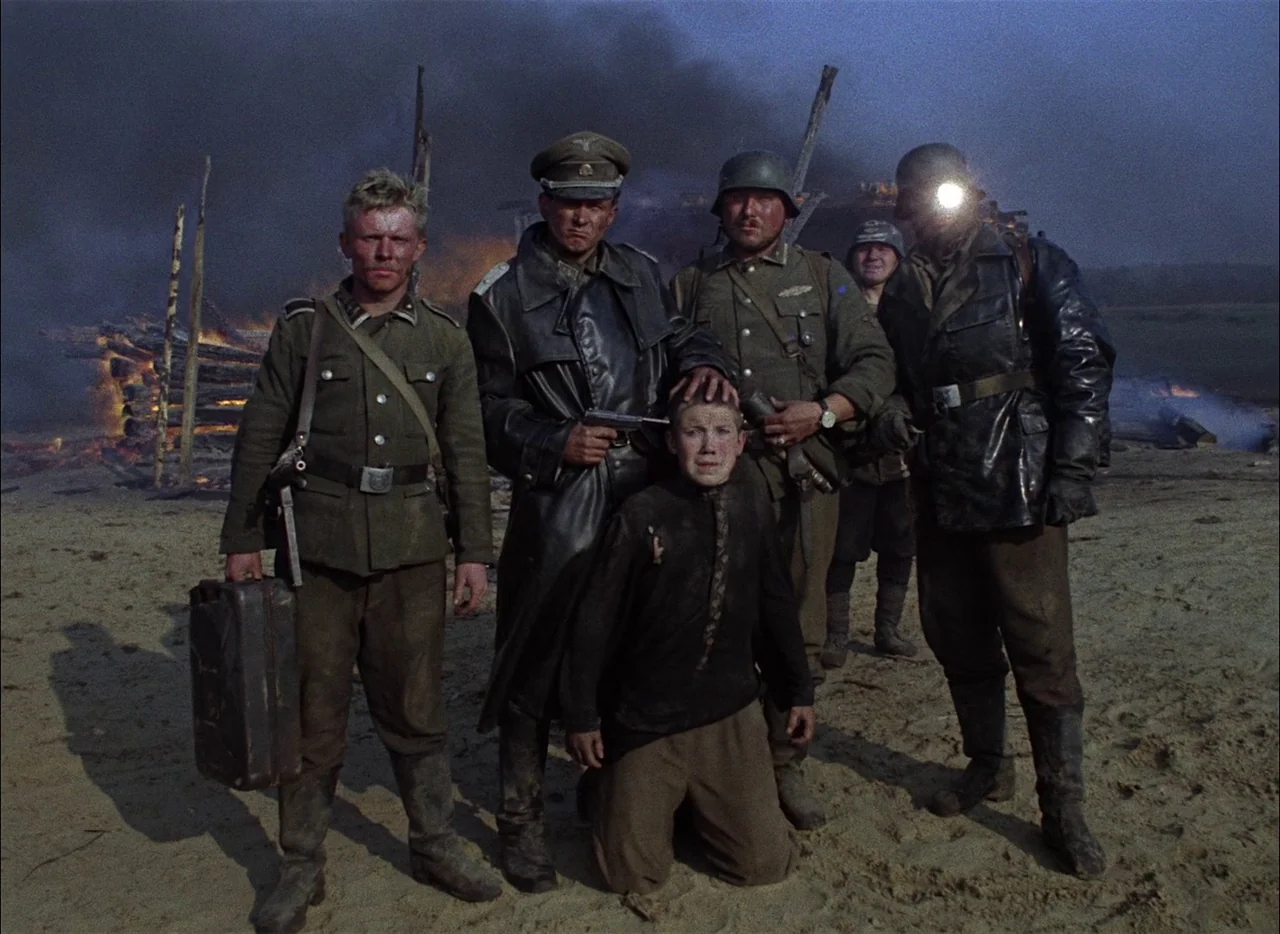 Come and See (1985) movie still: fullbody — A rural sandy clearing at twilight features a burning structure in the background. A group of four armed…
