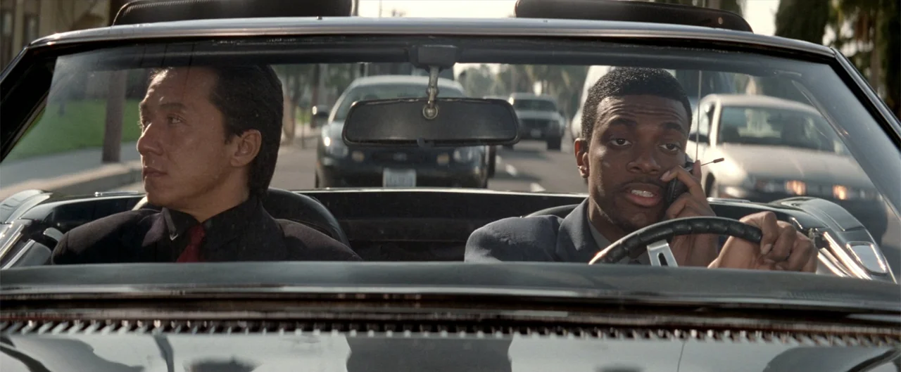 Rush Hour (1998) movie still: medium — A dark convertible travels along a suburban road during the bright daytime. Two men sit in the front seats…