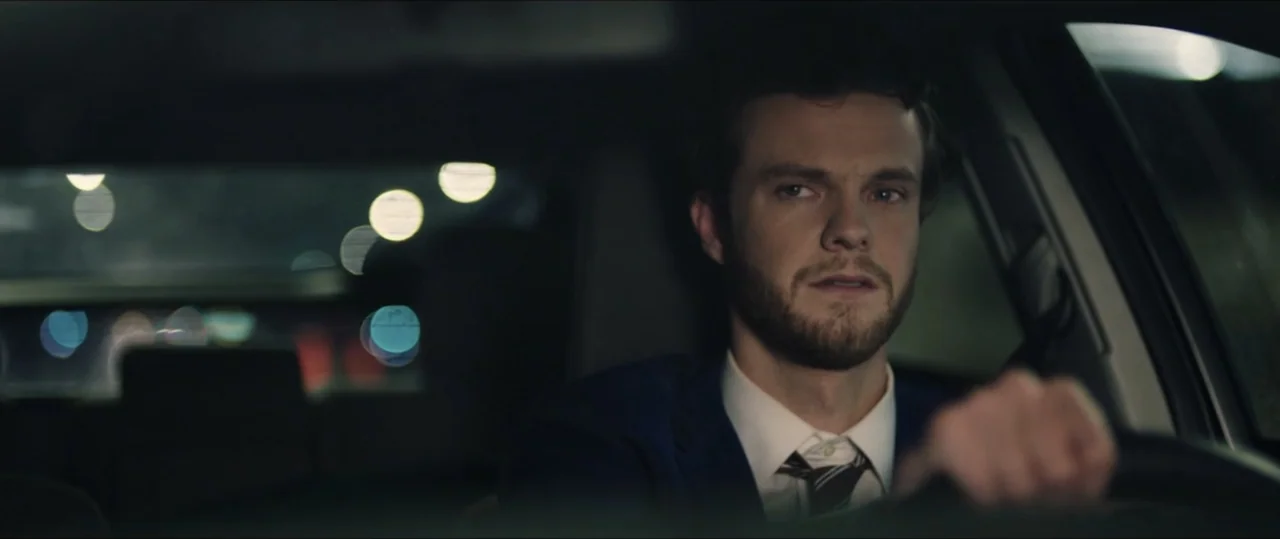 Plus One (2019) movie still: medium — A dark car interior at night shows blurred city lights through the rear window. A man in a suit drives the…