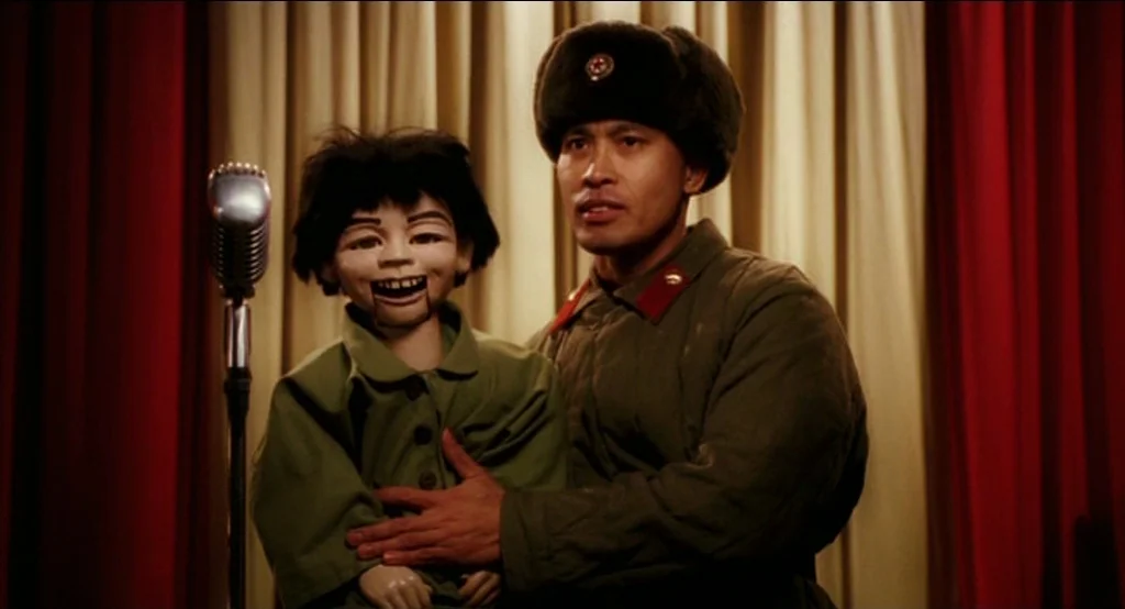 Big Fish (2003) movie still: medium — A man in a military uniform stands before a microphone holding a ventriloquist dummy. Behind them hang heavy…