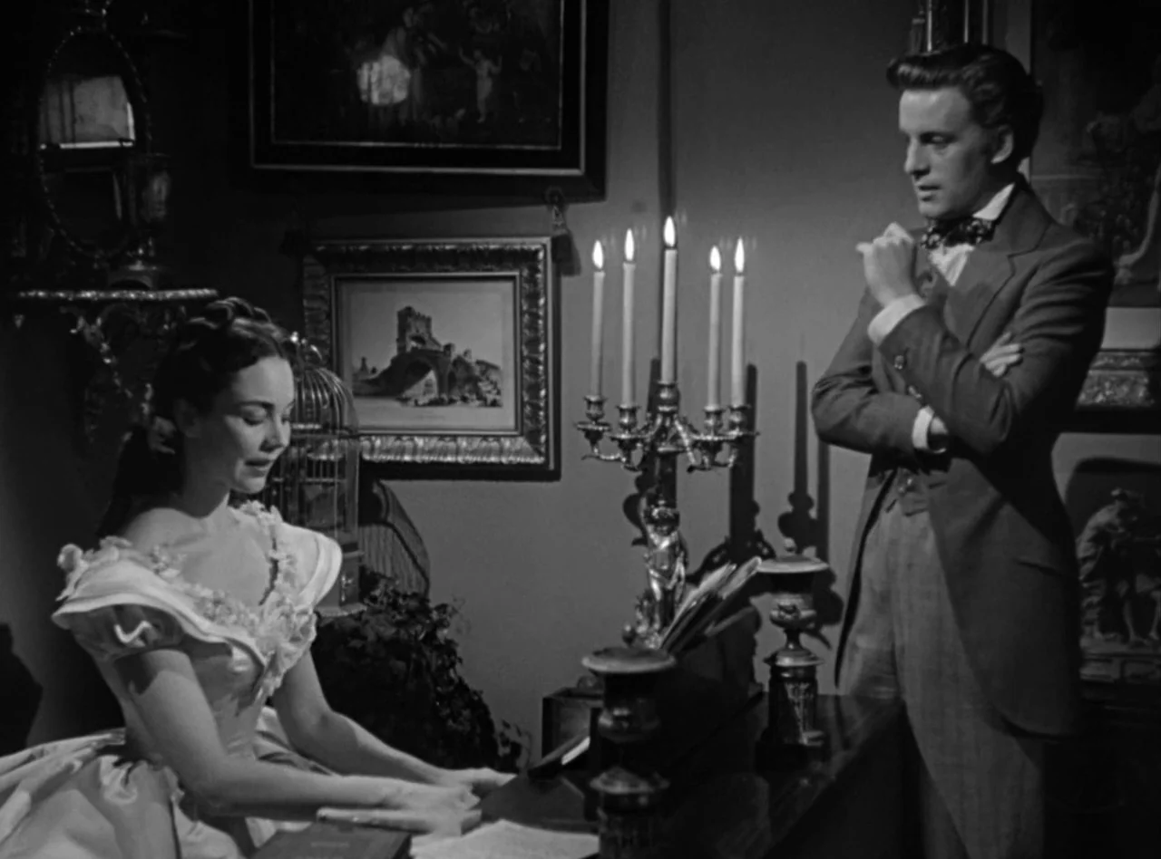 Madame Bovary (1949) movie still: medium — An interior parlor features ornate wall art and a silver candelabra on a piano. A young woman in a dress…
