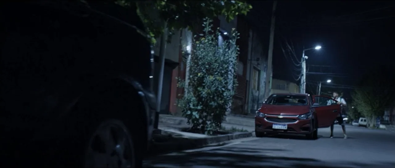 Gunman (2025) movie still: wide — A residential street at night features parked cars under artificial street lighting. A young man steps out of a…