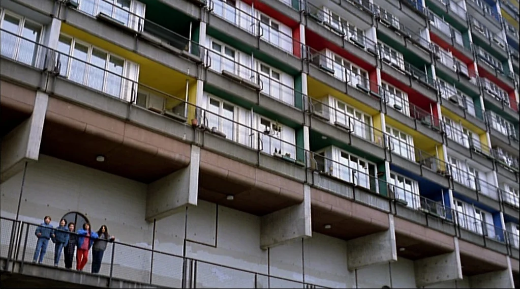 Wings Of Desire (1987) movie still: wide — A modern brutalist concrete apartment building features recessed balconies with bright red, yellow, and…