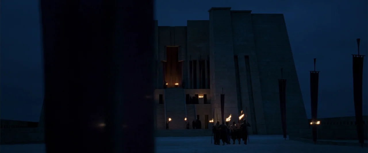 10,000 BC (2008) movie still: establishing — A massive stone temple with wide stairs stands beneath a deep blue night sky. A small group of…