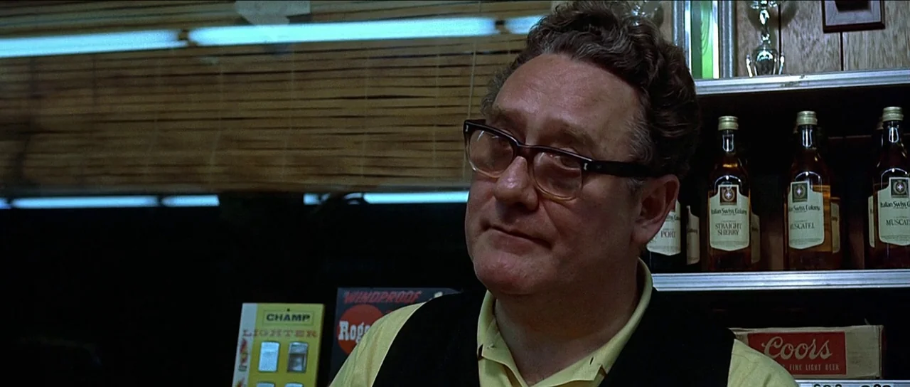 American Graffiti (1973) movie still: medium — A man stands in front of liquor store shelves featuring various glass bottles. The background…
