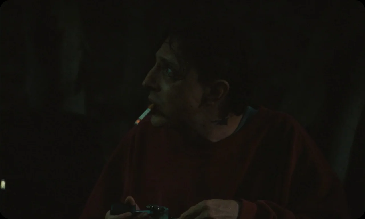 Vulcanizadora (2024) movie still: medium — A man sits alone in a dimly lit room with dark surroundings. He holds a small metallic object while a…