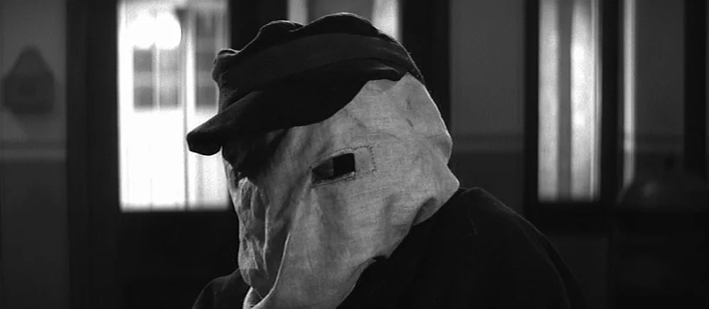 The Elephant Man (1980) movie still: closeup — A dimly lit interior room features a doorway with light leaking through the frame. A person wearing a…