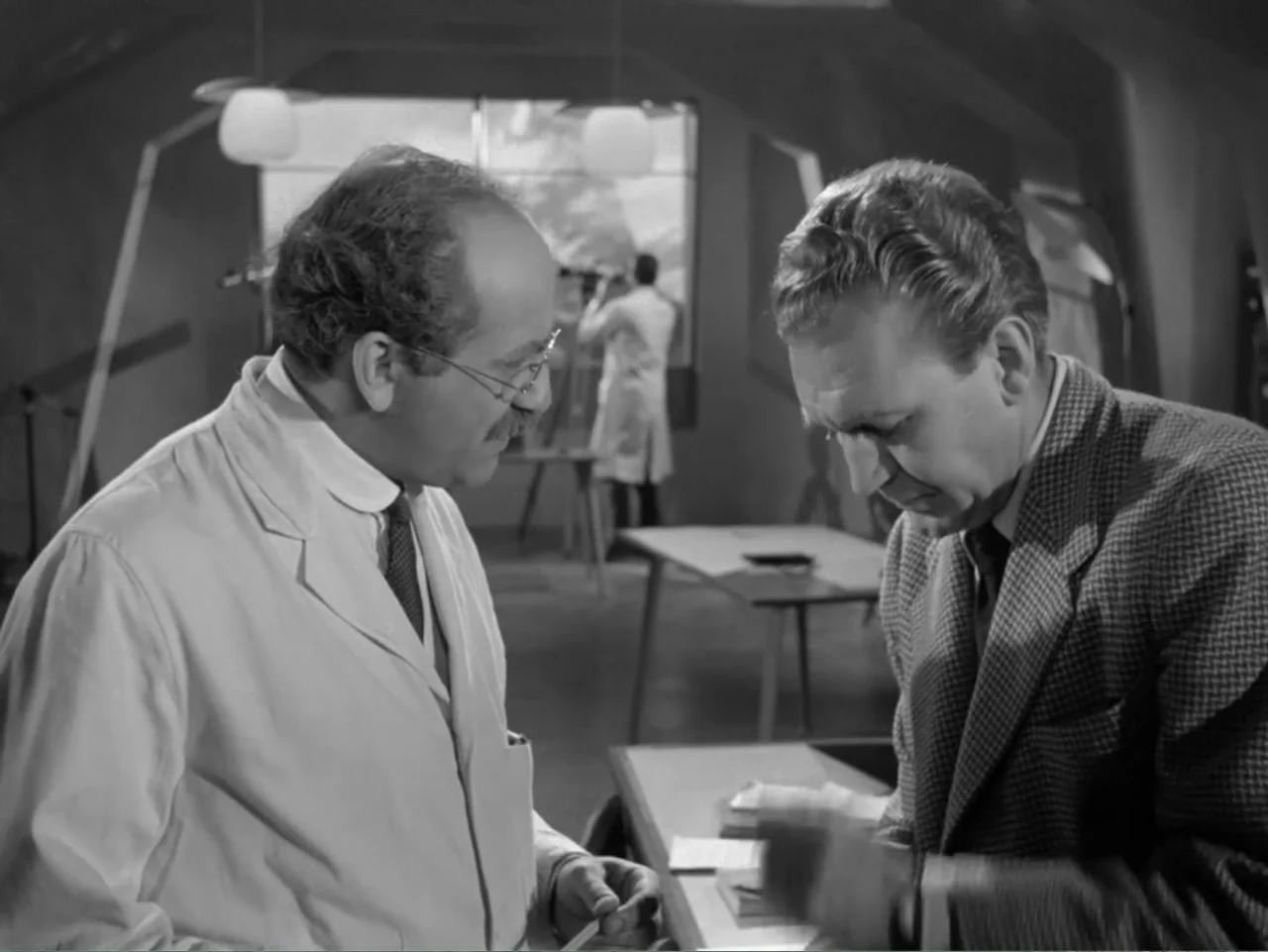The Crawling Eye (1958) movie still: medium — A medical laboratory with tables and scientific equipment in the background. Two men engage in…