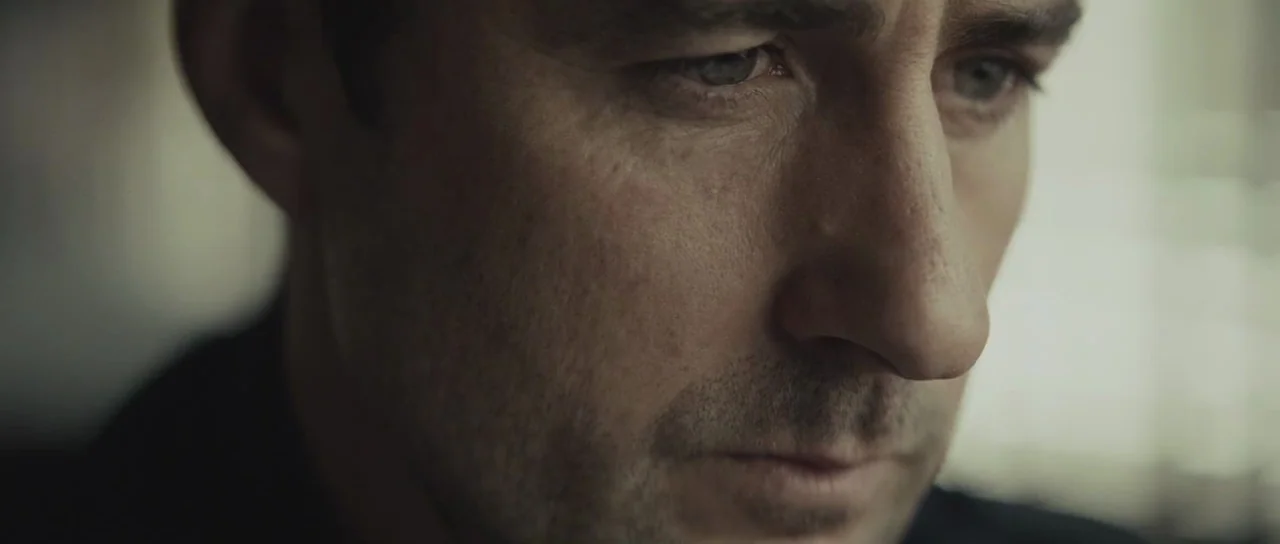 Meadowland (2015) movie still: closeup — A close-up shot of a man's face in a dimly lit indoor environment. The man looks downward while soft…