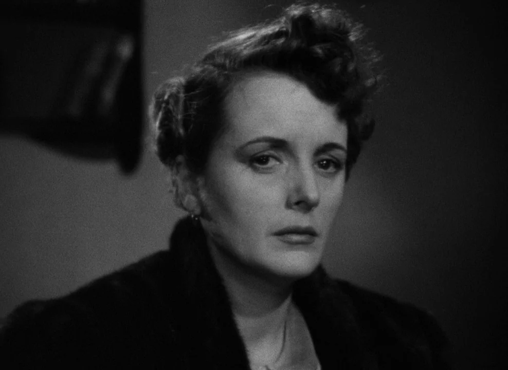 The Maltese Falcon (1941) movie still: closeup — A woman stands in a dimly lit room with plain walls. She faces the lens directly while wearing a…