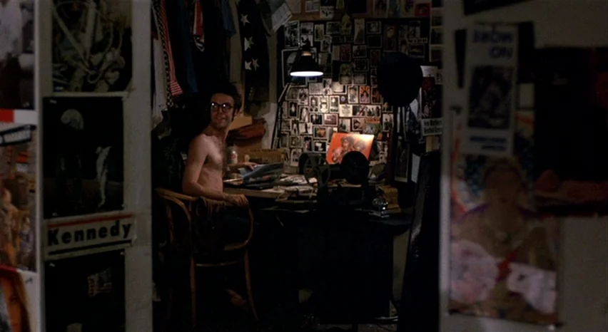 A Safe Place (1971) movie still: medium — A cramped, dark workspace filled with walls of posters and miscellaneous clippings. A shirtless man sits…