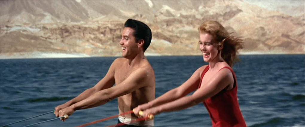 Viva Las Vegas (1964) movie still: medium — Two people water ski across a lake with dry, rocky hills in the background. A man and a woman hold onto…