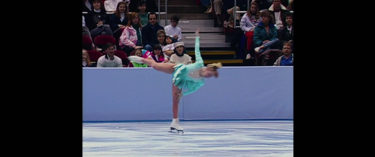 I, Tonya (2017) movie still: fullbody — A figure skater performs on an ice rink in front of a seated audience. The skater is executing a balanced…