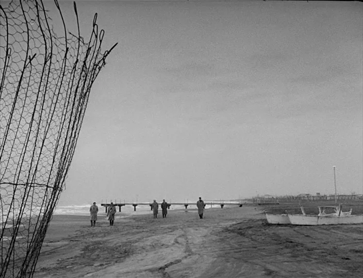 I Vitelloni (1953) movie still: wide — A desolate, overcast beach scene with a wooden pier extending into the water. Five men in long coats walk…