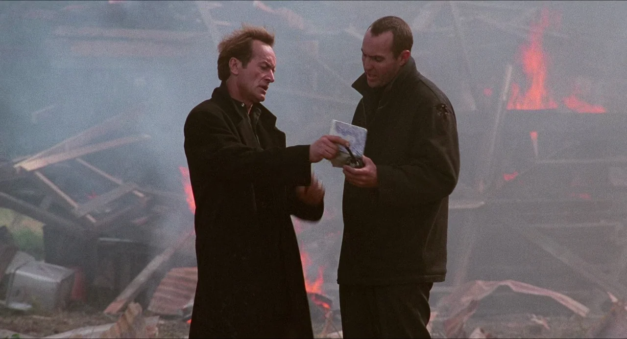 Hard Target (1993) movie still: medium — A pile of burning wooden debris and wreckage fills the background with rising smoke. Two men stand in the…