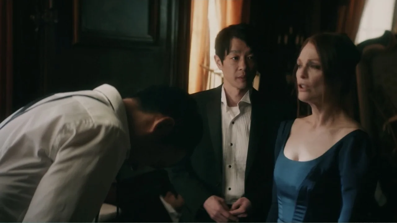 Bel Canto (2018) movie still: medium — A dimly lit room with wood paneling and window curtains in the background. A man bows deeply to a woman and…