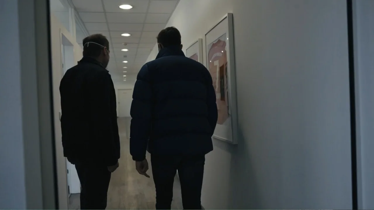 Federico Chiesa - Back on Track (2023) movie still: fullbody — A long brightly lit hallway with white walls and recessed ceiling lights. Two men…
